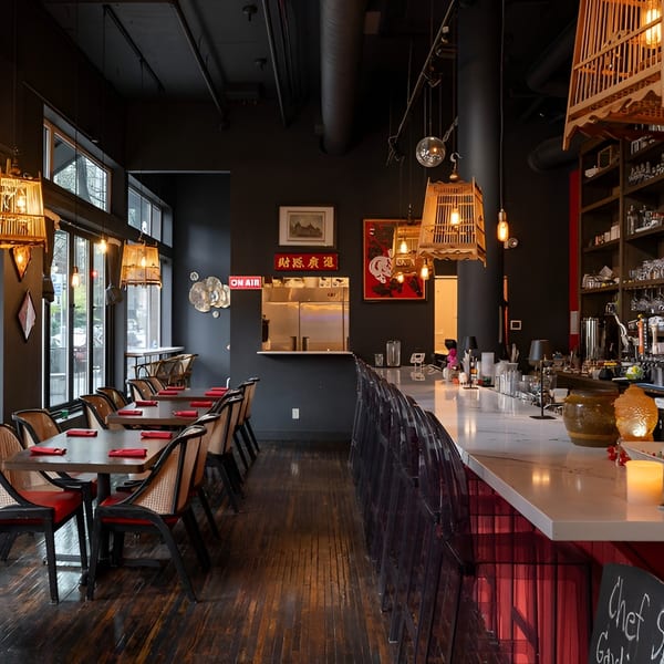 Restaurant interior with dark walls, wooden tables, wicker chairs, a long bar, and decorative lighting.