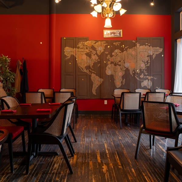 Restaurant dining room with red walls, dark wood floors, cane-back chairs, and a world map mural.