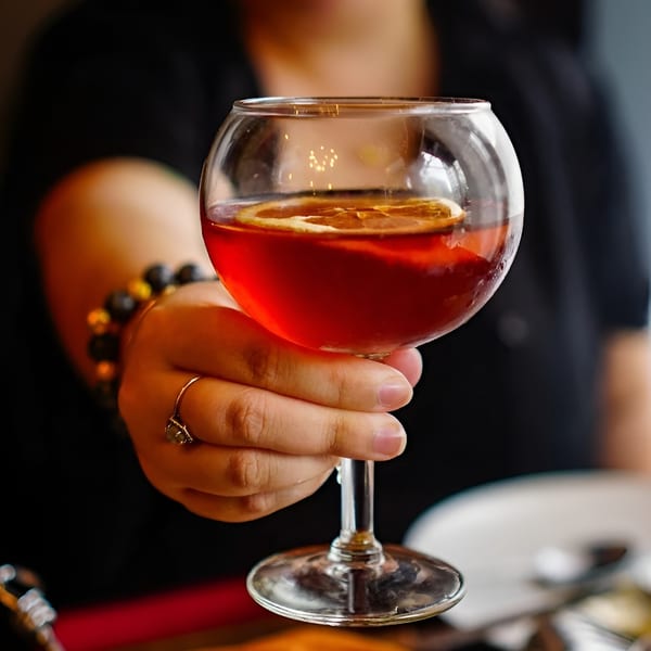A person holding a glass of red cocktail with an orange slice, with food in the foreground.