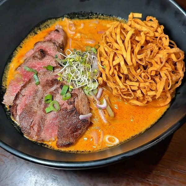Khao Soi with sliced beef, crispy noodles, sprouts, and green onions in a black bowl.