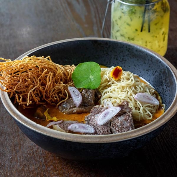 Khao Soi with beef, crispy noodles, fresh noodles, shallots, and a green leaf in a dark bowl.