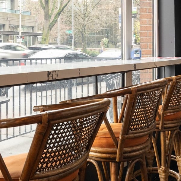 Rattan bar stools with orange cushions line a window counter overlooking a city street.