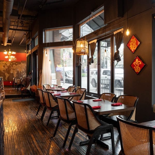 Interior of a restaurant with a long bar, dining tables, and large windows looking out onto a street.