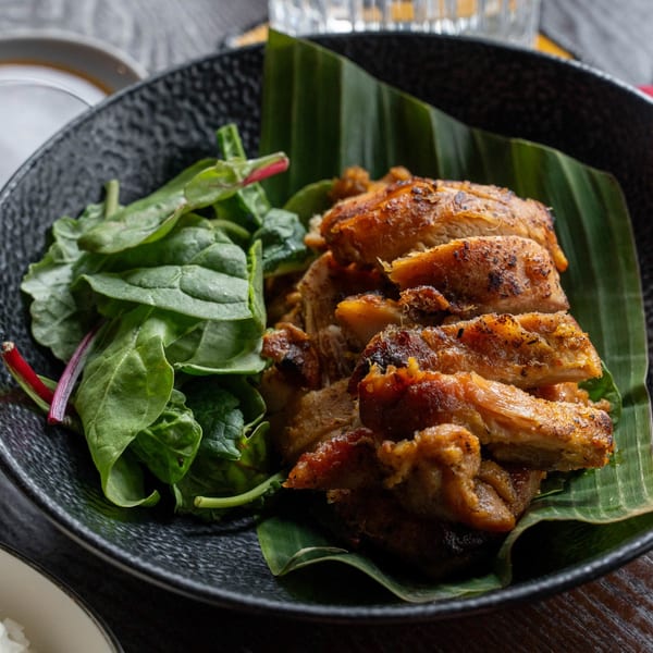 Grilled chicken slices on a banana leaf with a side of fresh spinach in a black bowl.